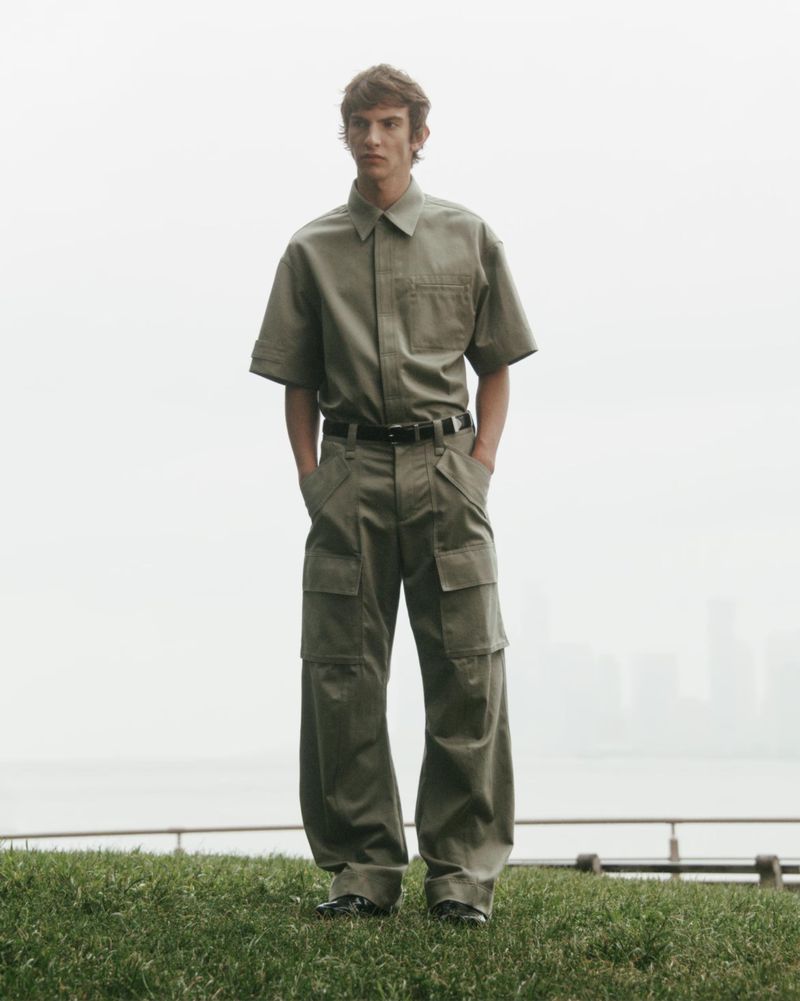 Fashion shot of a young adult man in a short-sleeved khaki shirt and khaki cargo pants, posing on a grass hill with a cityscape background.