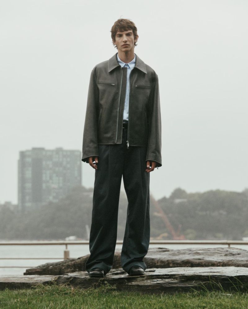 Adult male fashion model poses in a grey jacket and wide-leg pants in an outdoor environment with a city and greenery visible.