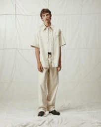 Model standing against a white textured backdrop wearing a cream button-up shirt and matching trousers.