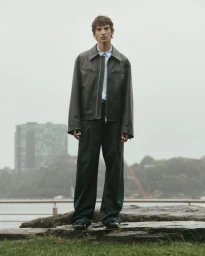 Model wearing a dark leather jacket and trousers standing outdoors against an overcast waterfront background.