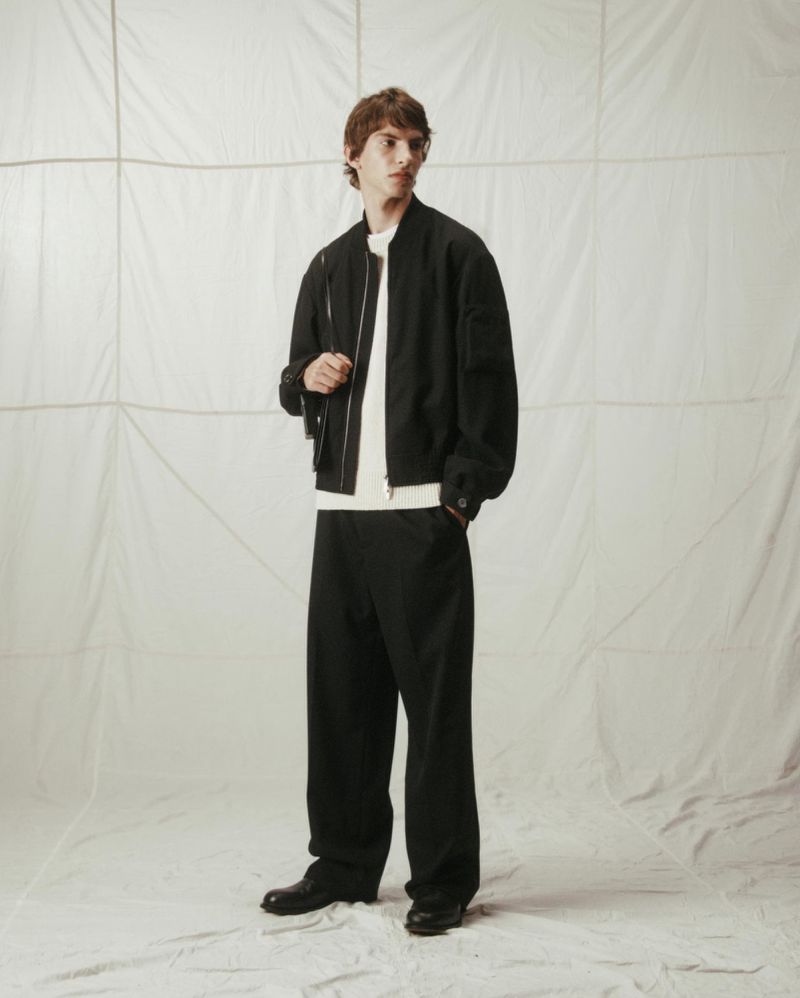 Model wearing a black zip-up bomber jacket and black trousers against a textured white sail backdrop.