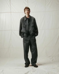 Full-length portrait of a male model wearing a dark grey jacket and trousers against a textured white backdrop.