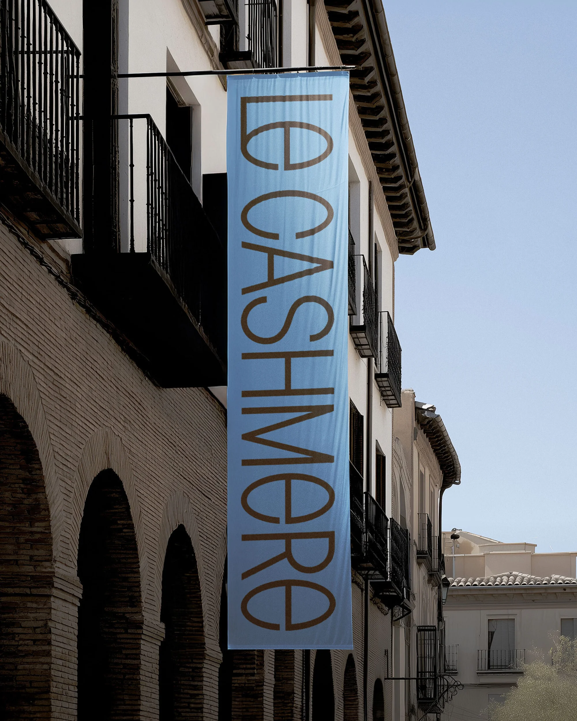 Vertical banner for Le Cashmere brand hangs on building with white walls, arched doorway, and black wrought iron balconies, daytime.