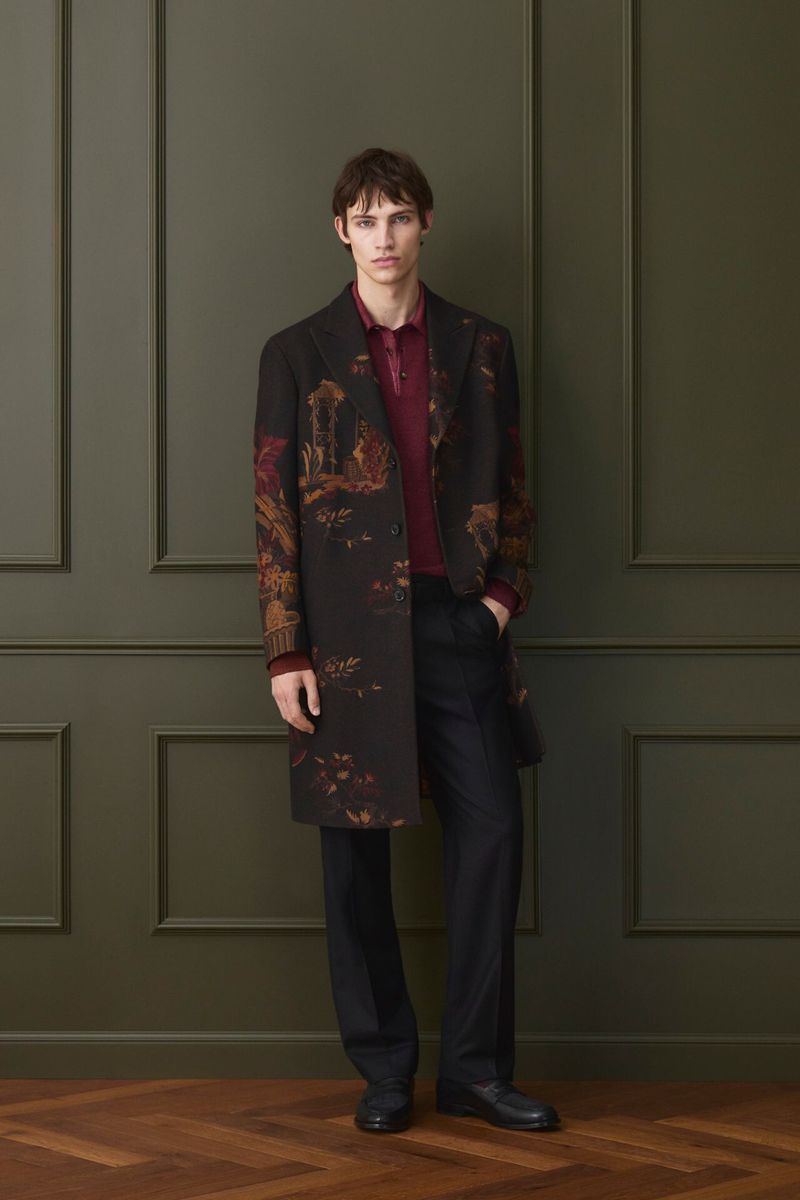 A young adult male stands in a fashionable patterned coat, burgundy polo shirt, and black pants in front of a dark green wall.