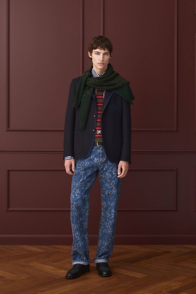 Full-length shot of a male fashion model in a dark blazer, printed denim jeans, and a green scarf, posing against a paneled wall.