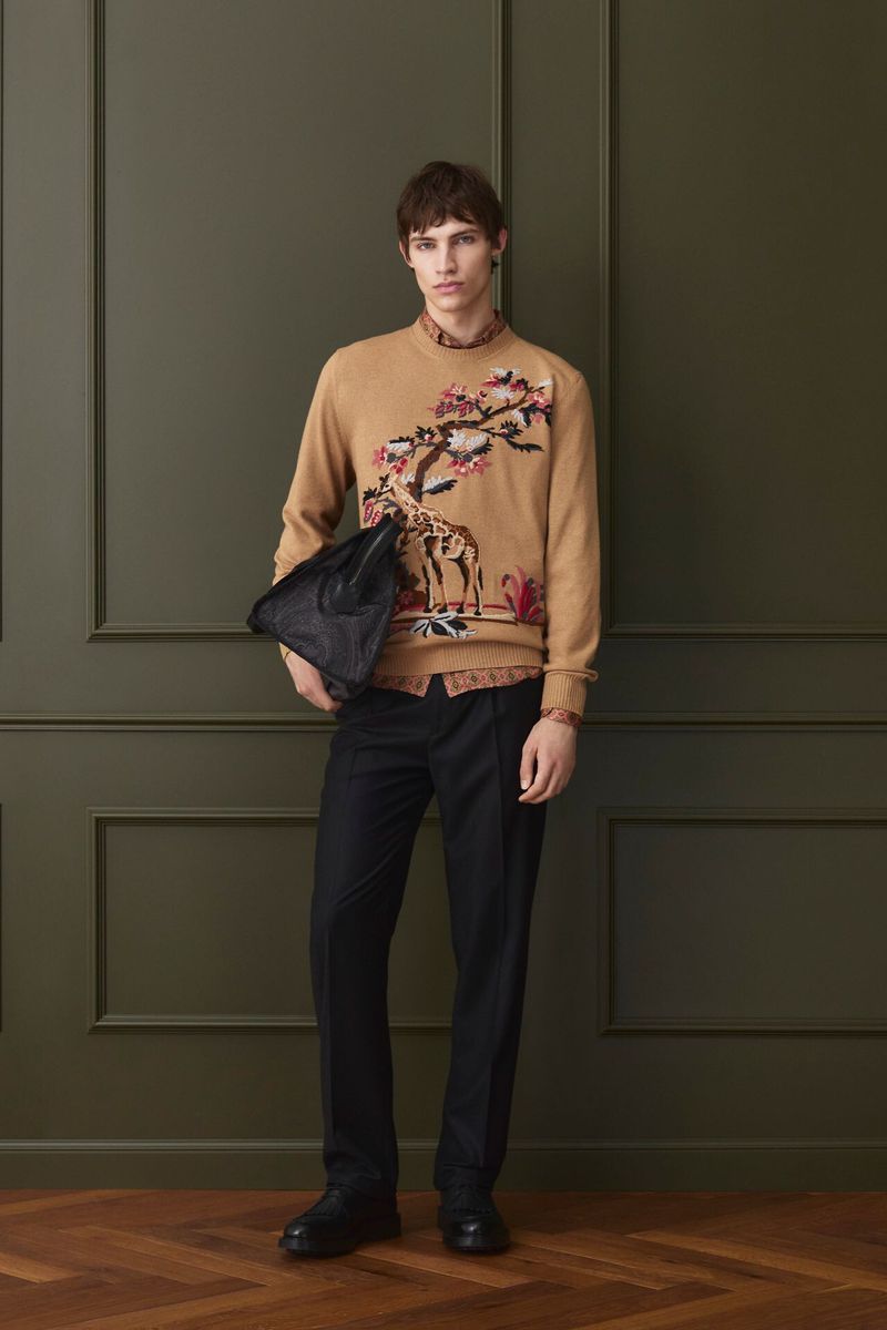 Male model wearing a camel sweater with embroidered animal motif and dark trousers holding a bag against green paneling