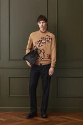 Male model wearing a camel sweater with embroidered animal motif and dark trousers holding a bag against green paneling