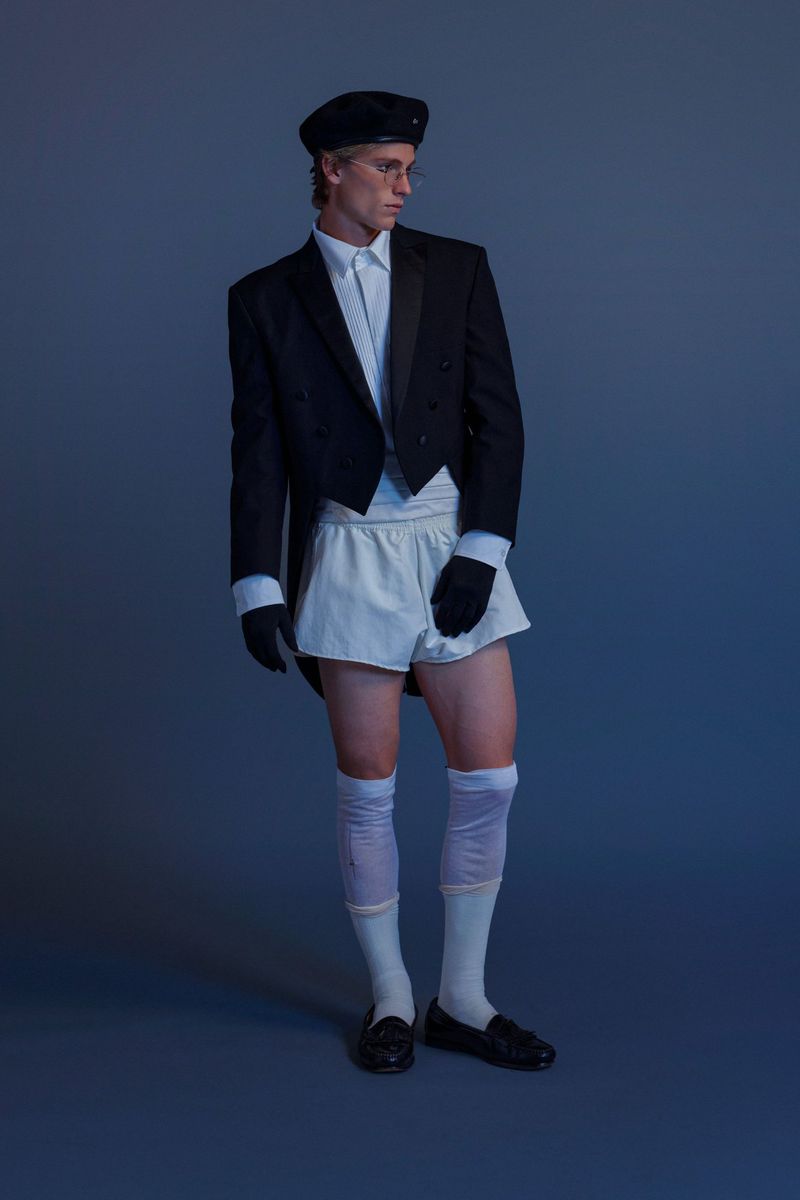 Man in tuxedo jacket, white shorts, opera gloves, beret, knee-high socks and loafers posing in studio with a blue backdrop.