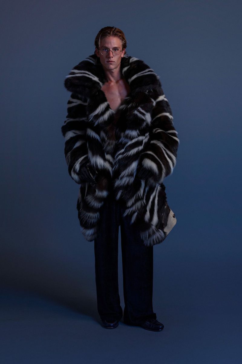Fashion shot of a man with blond hair and glasses wearing a black and white striped fur coat and black pants against a blue backdrop.