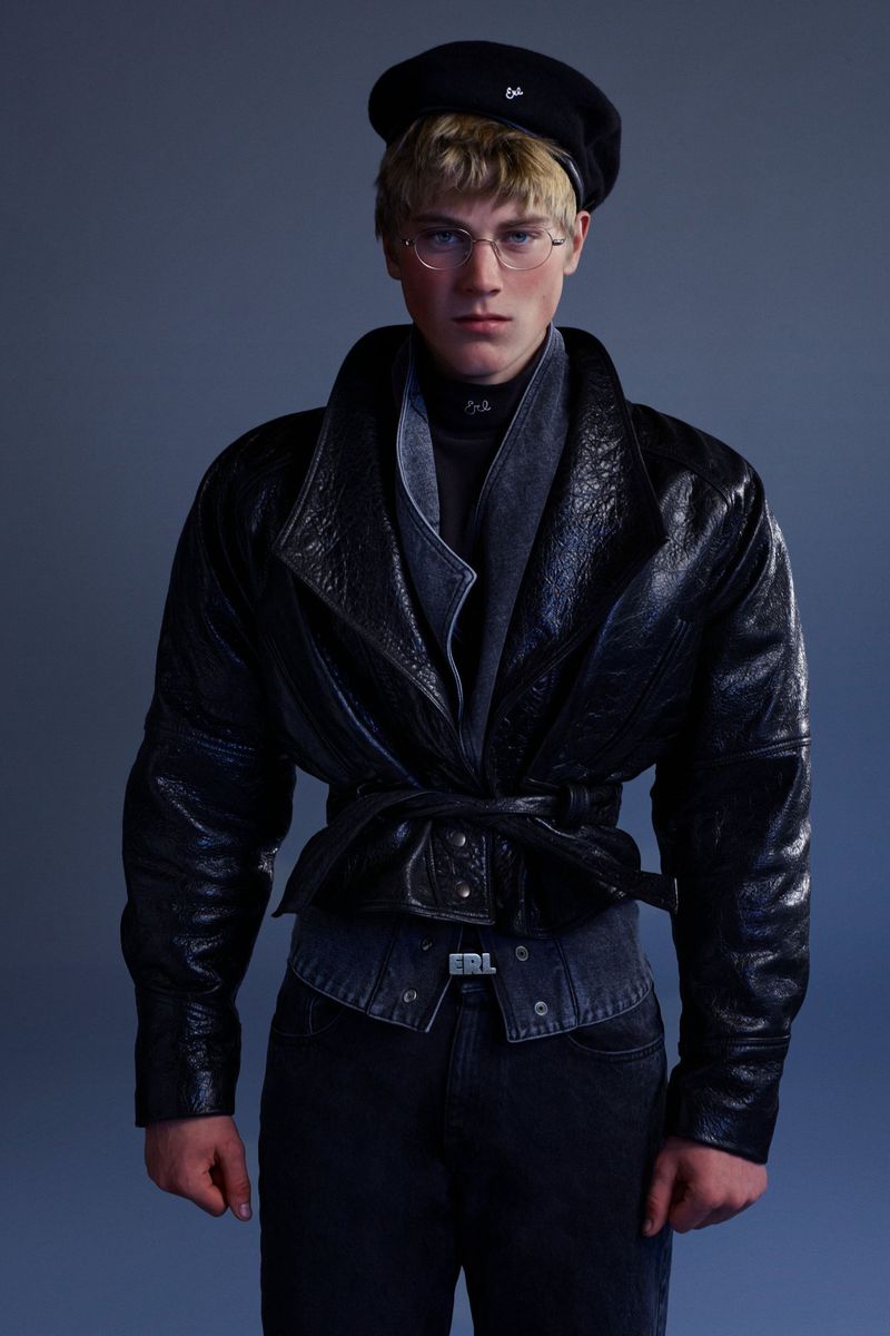 Adult male in a black leather jacket, black pants, and a black beret poses in a studio. He wears clear framed glasses.