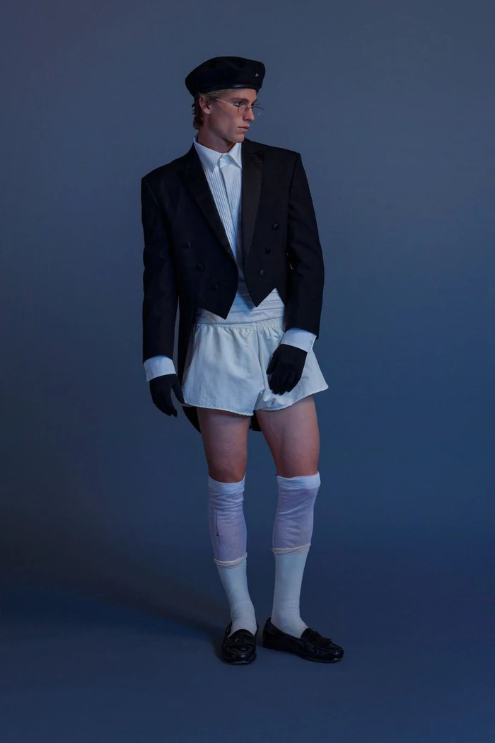 Model wearing a black tuxedo jacket, white shorts, beret, and knee-high socks against a dark grey studio background.