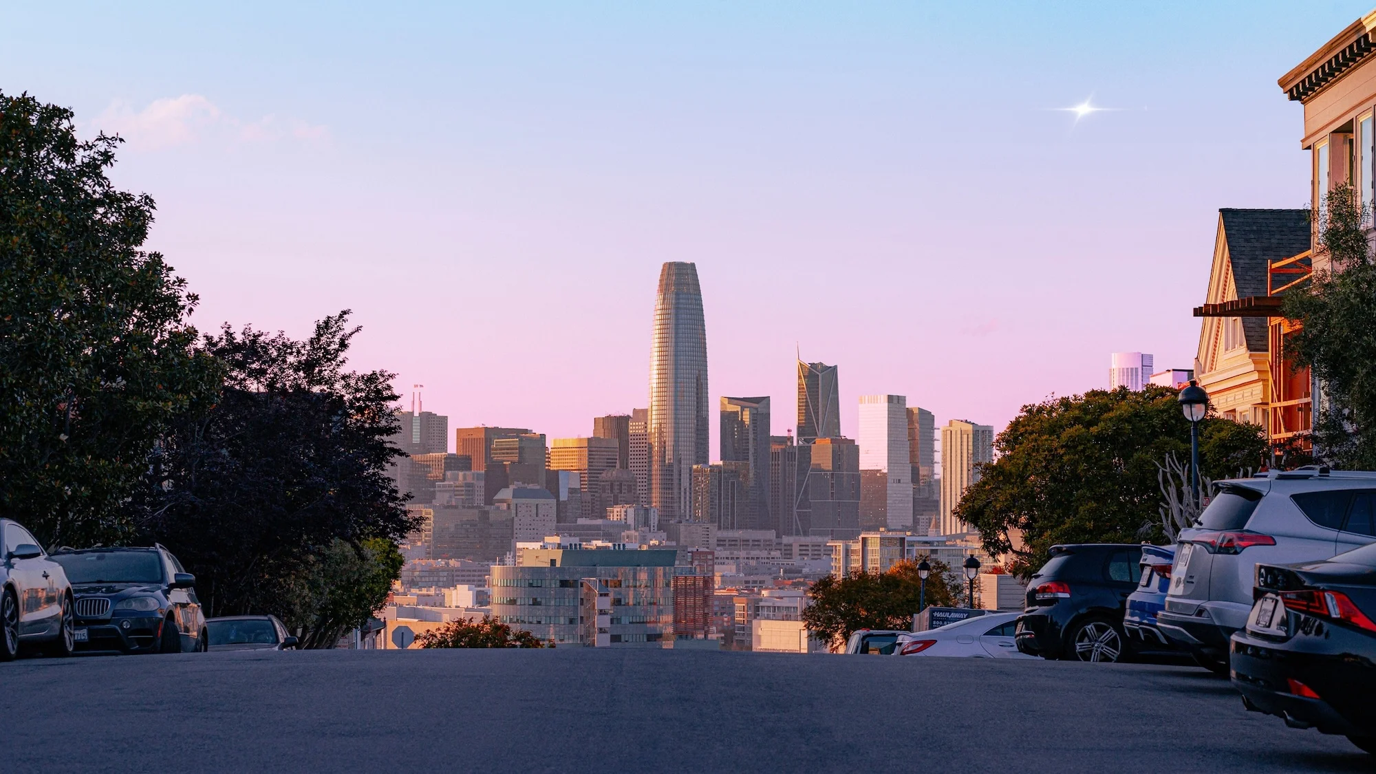 San Francisco skyline at sunset featuring the Salesforce Tower, seen from a street with parked cars and trees, creating a serene urban view.