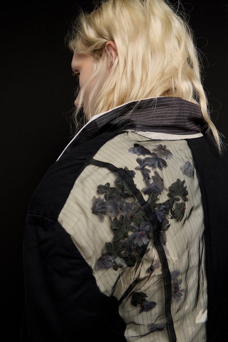 Blonde haired adult woman wearing black and sheer floral patterned jacket. Dark background. Fashion shot, back view.