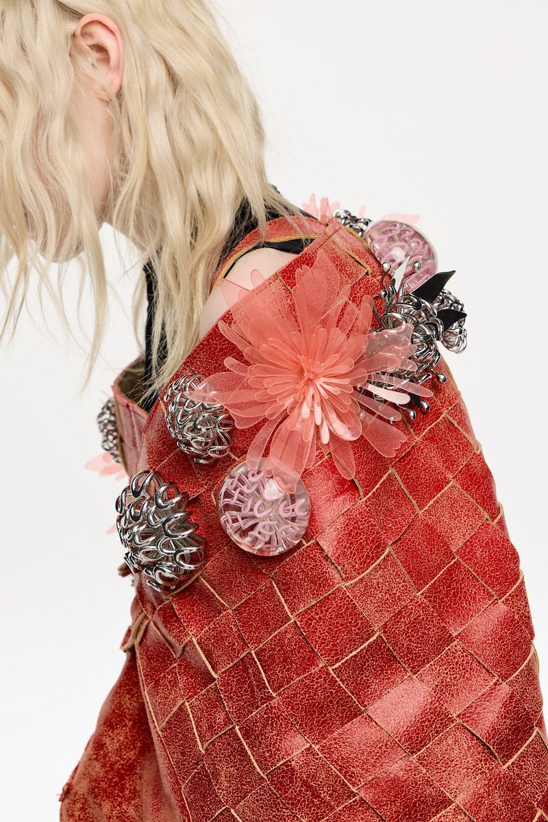 Woven red garment with floral and metallic embellishments, worn by a blonde person against a clean white background. Fashion-forward style.