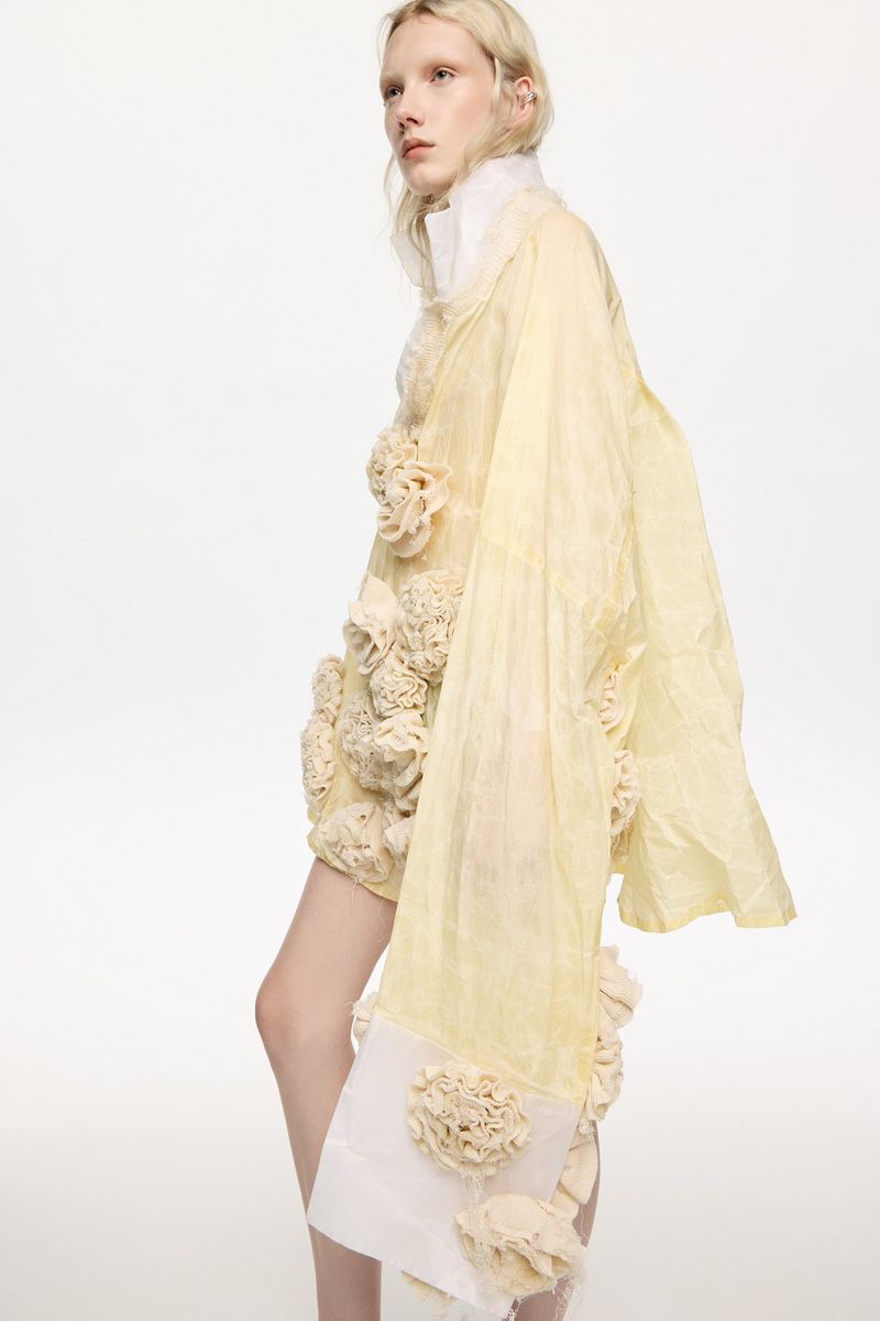 Fashion shot of a female model in a pale yellow floral garment against a white background, showcasing textile art and couture design.