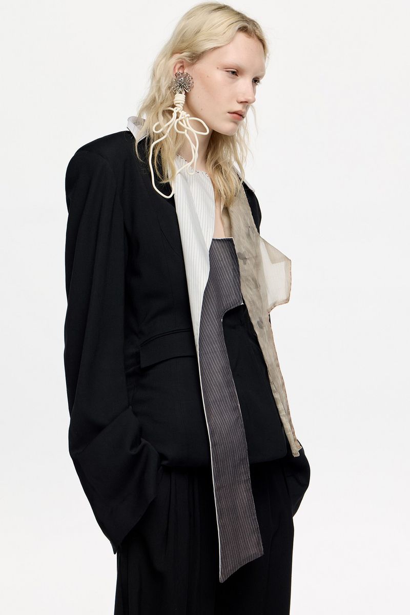Fashion portrait of a young, light-skinned woman in an oversized black blazer, striped top, large decorative earring, and dark pants.