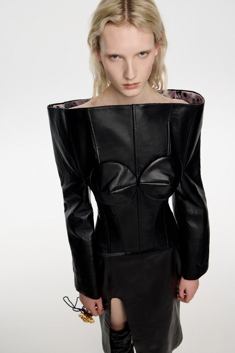 A pale-skinned woman in an avant-garde black leather outfit poses against a white background in a fashion studio, showcasing a unique design.