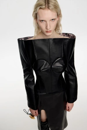 A pale-skinned woman in an avant-garde black leather outfit poses against a white background in a fashion studio, showcasing a unique design.