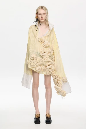 Model wearing a pale yellow asymmetric upcycled garment adorned with fabric rosettes and black platform shoes against a white background