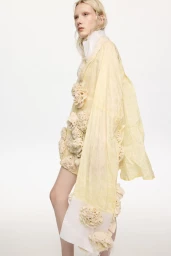 Model wearing a pale yellow textured garment adorned with fabric rosettes against a plain white studio background.