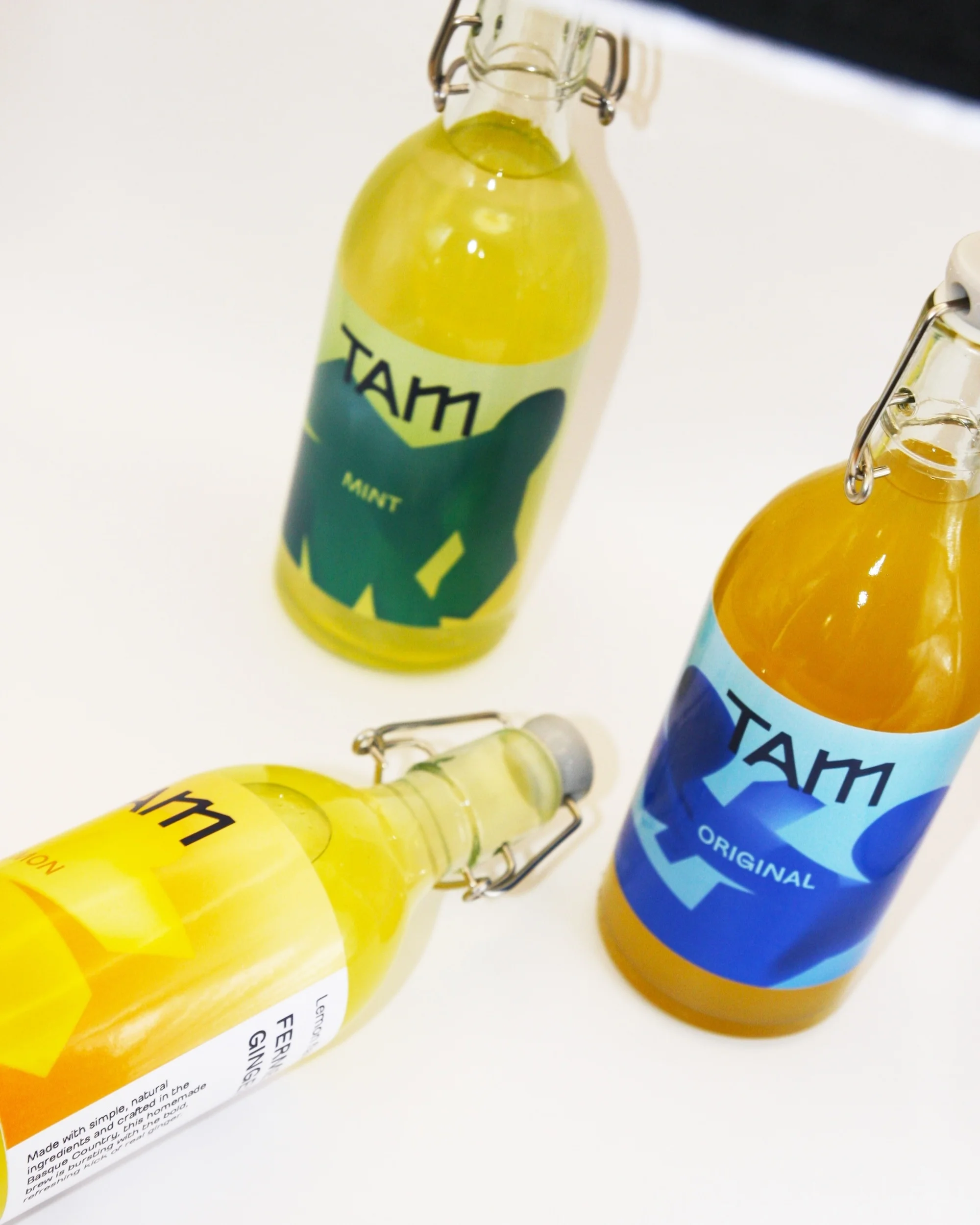 Three glass bottles of artisanal beverages with labels 'TAM' in an indoor setting on a white surface, showcasing a minimalist aesthetic.