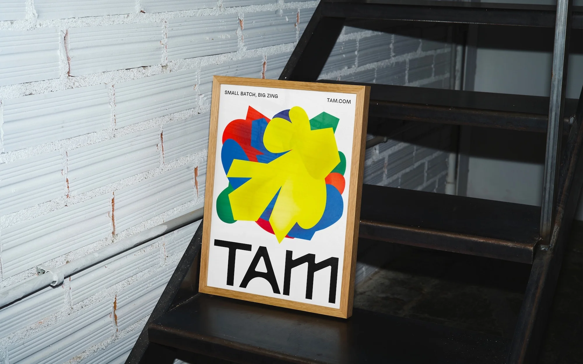 Framed poster with abstract colorful graphics and text, 'TAM,' leaning against a white brick wall on dark metal steps, minimalist.