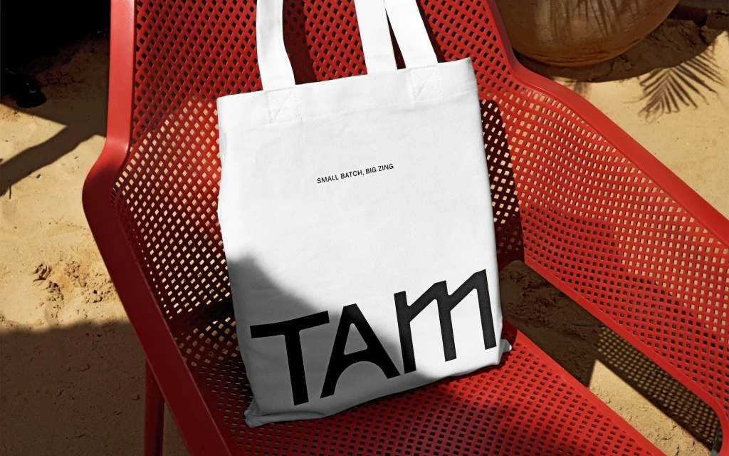 White canvas tote bag with black text on a red mesh chair in an outdoor setting with sand and a coconut in the background.