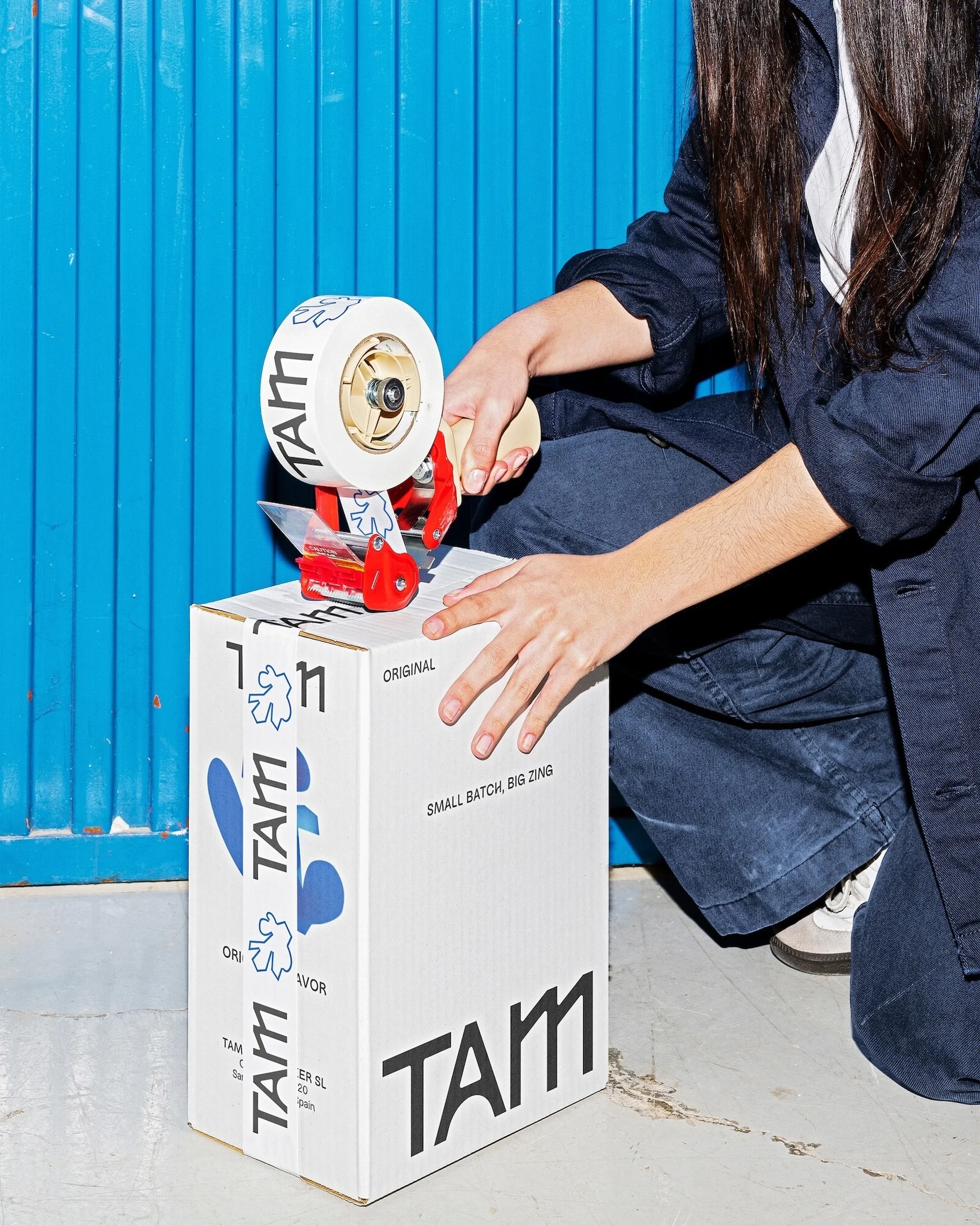 Adult packs a white cardboard box with a tape dispenser. The box features a company logo. A blue wall is in the background.