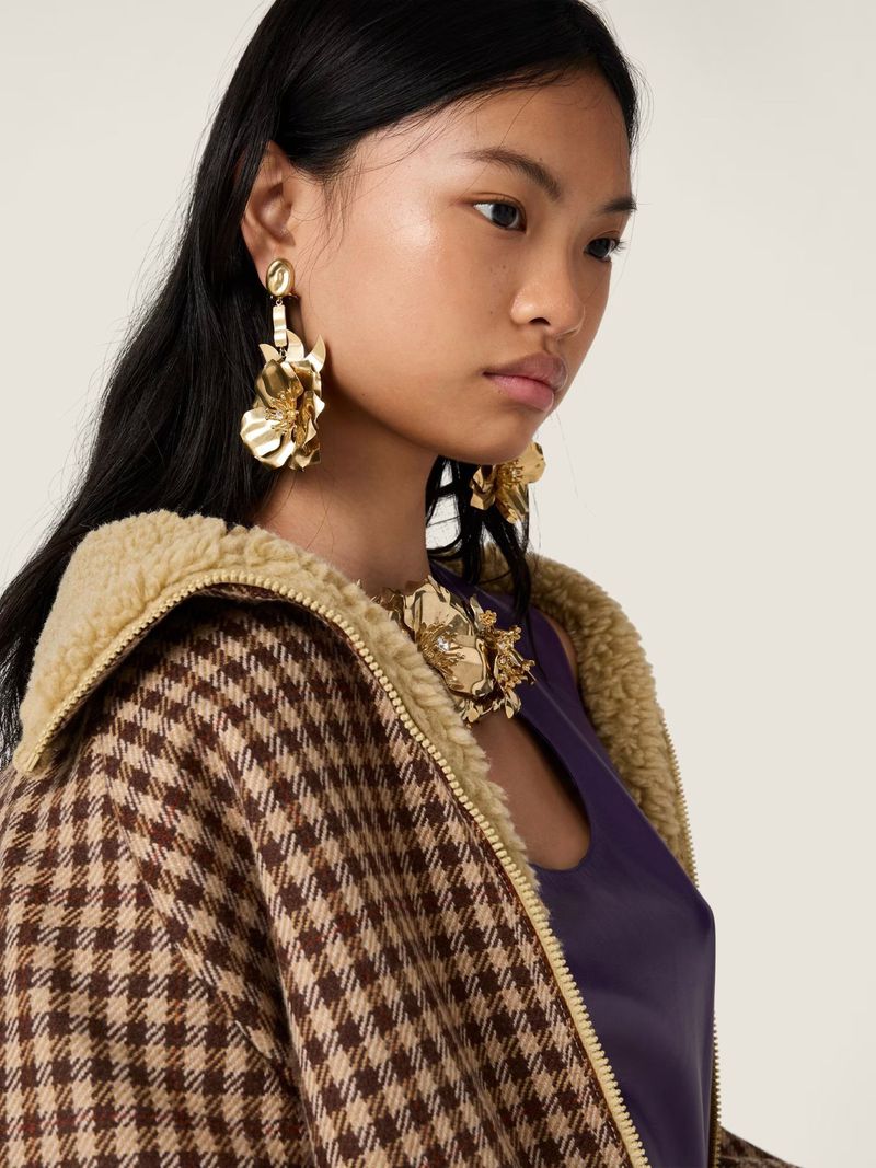 Close-up of a woman wearing gold floral earrings and necklace, a brown and beige houndstooth jacket, and a purple top, posing indoors.