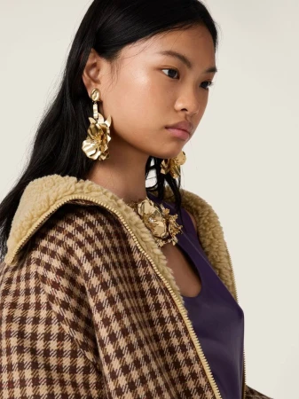 Close-up of a woman wearing gold floral earrings and necklace, a brown and beige houndstooth jacket, and a purple top, posing indoors.