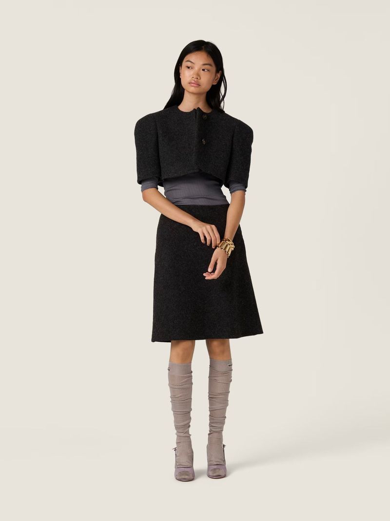 Elegant woman in a gray wool skirt suit with a cropped jacket, knee-high boots, and gold bracelet posing in a neutral studio setting.