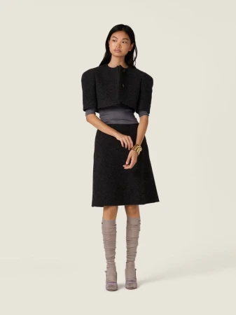 Elegant woman in a gray wool skirt suit with a cropped jacket, knee-high boots, and gold bracelet posing in a neutral studio setting.