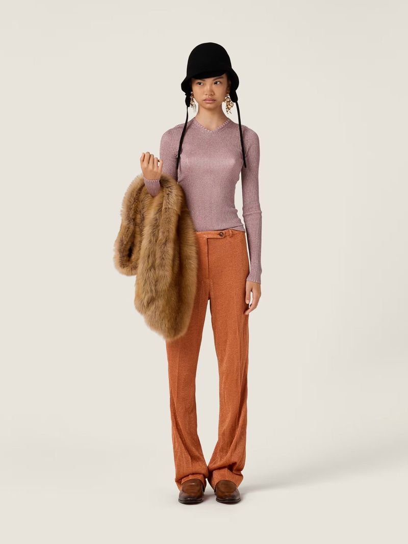 Full-length fashion shot: Asian woman in a lilac top and orange pants poses with a fur jacket against a solid off-white backdrop.