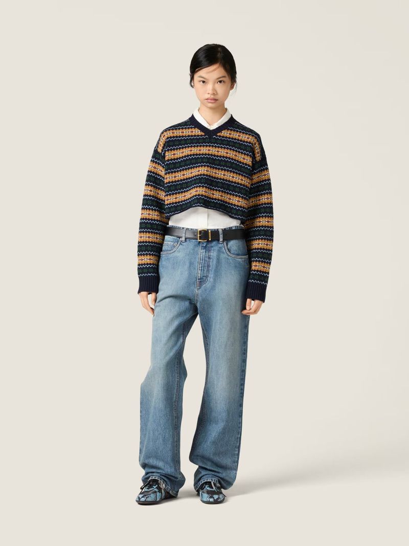 An Asian woman wearing a cropped sweater, button-down shirt, wide-leg jeans, and chunky shoes stands in a brightly lit neutral studio.