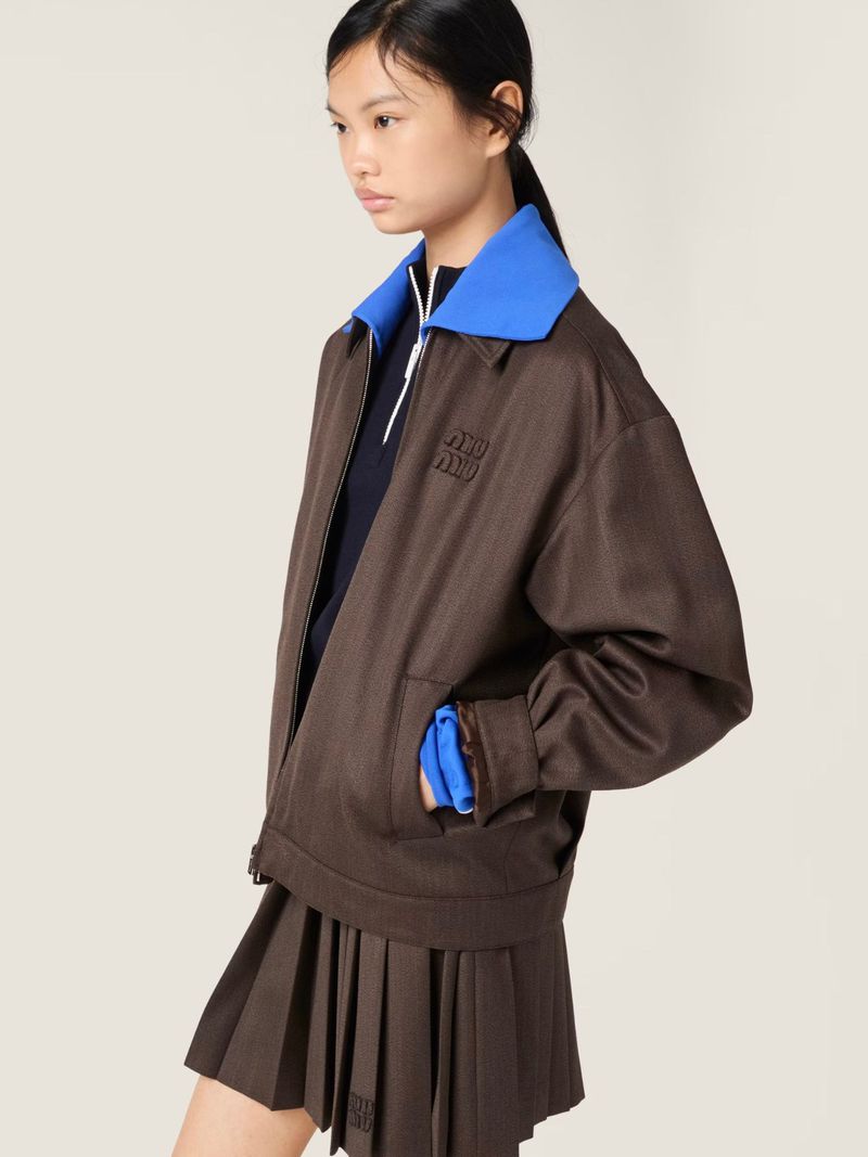 Young Asian woman posing in a brown jacket and pleated skirt with the Miu Miu logo; studio shot on a cream background for a fashion layout.