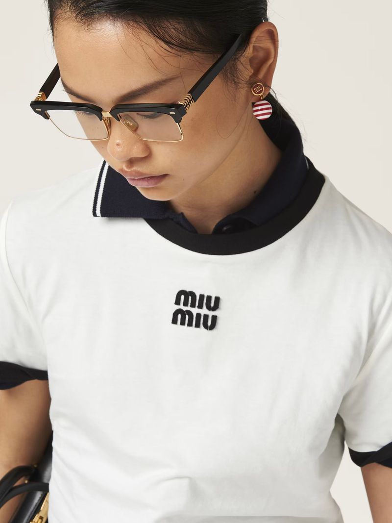 A woman in eyeglasses and a white MIU MIU logo t-shirt against an off-white backdrop, offering a clean, fashion-forward aesthetic.