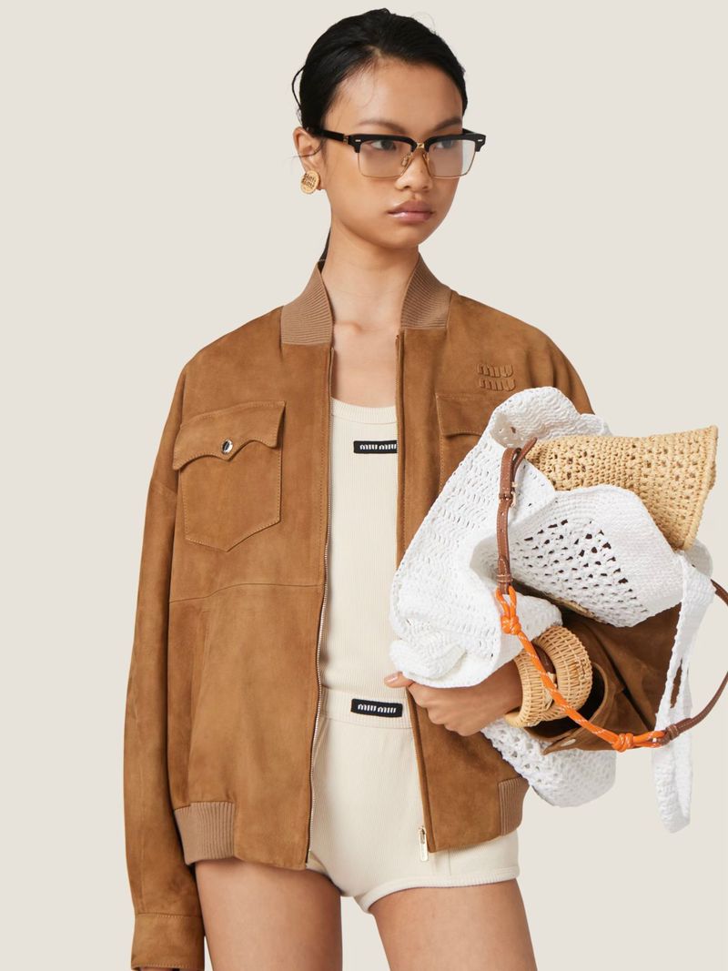 Woman in brown suede jacket, white top and shorts holding straw bags in studio setting. Fashion, beauty and lifestyle photography.
