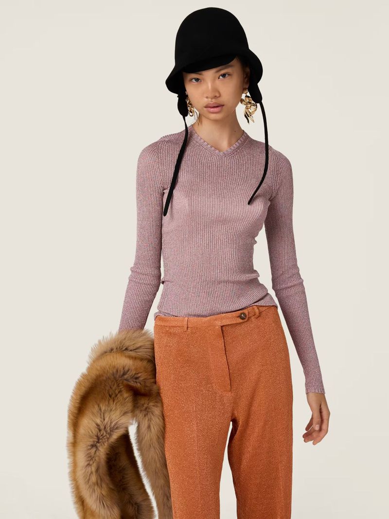 Indoor fashion shot of an Asian female model in a lilac top, orange pants, and a black hat, holding a faux fur stole, against a white backdrop.