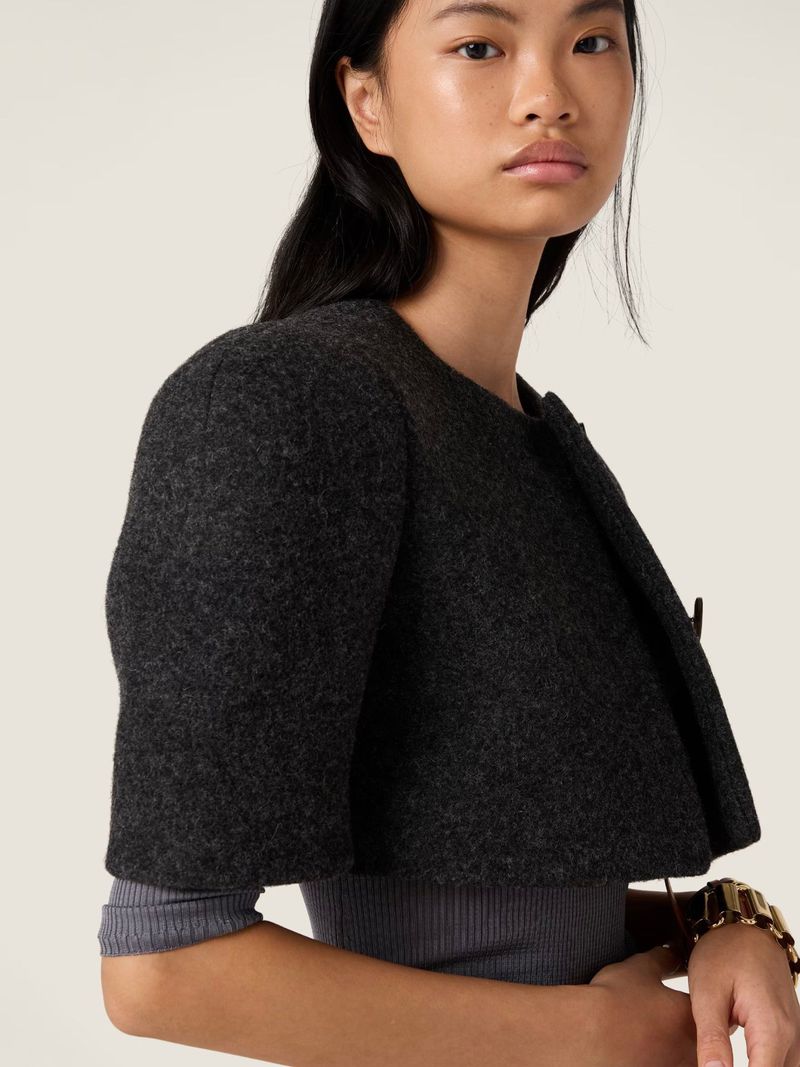 Close-up of an Asian woman wearing a cropped dark grey wool jacket and a grey top against an off-white backdrop in a studio shot.