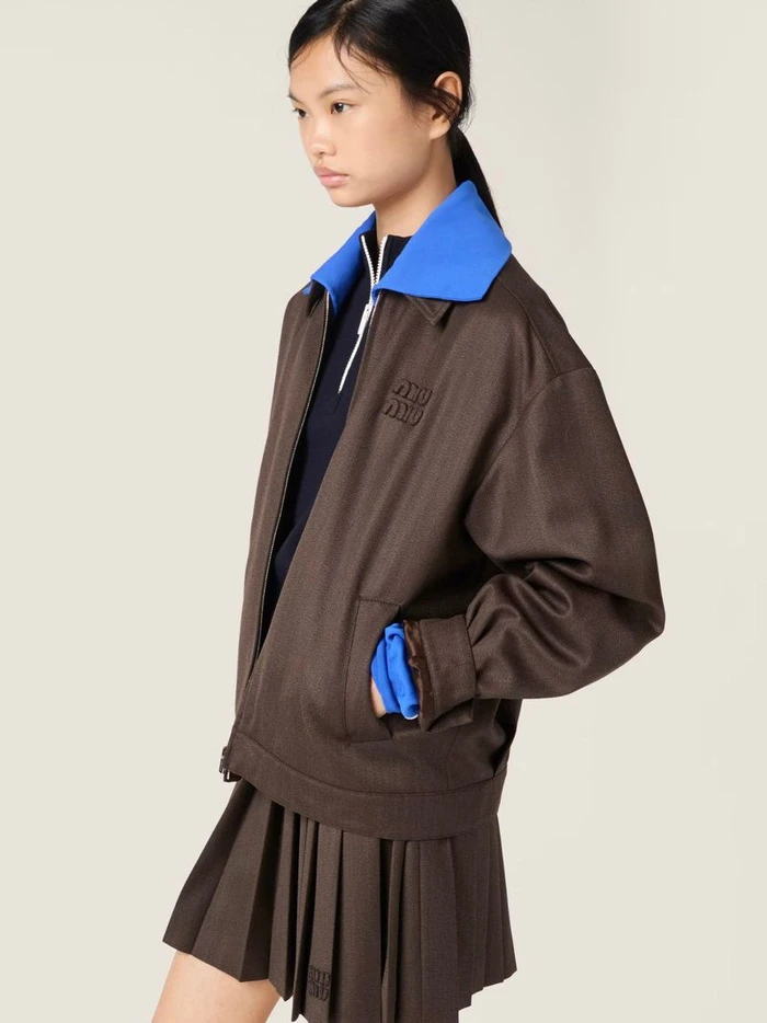 Model Yue Gu wearing a brown jacket with a blue collar and pleated skirt against a plain seamless background.