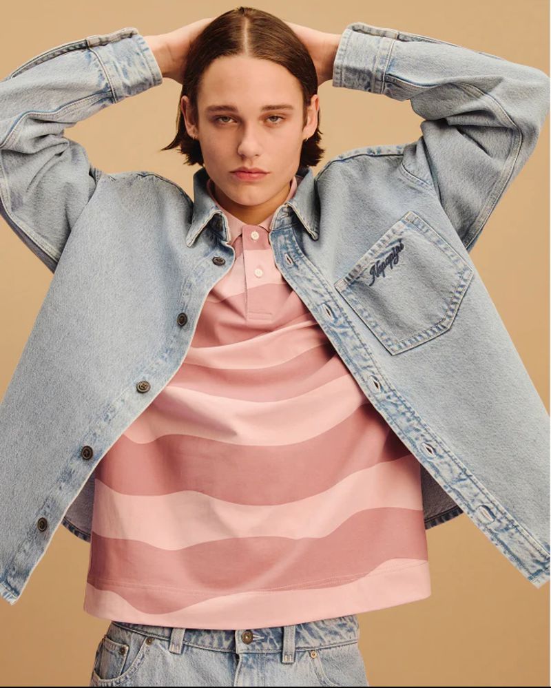 Light denim jacket and striped pink polo shirt worn by a young adult male against a neutral backdrop in a studio-style portrait.