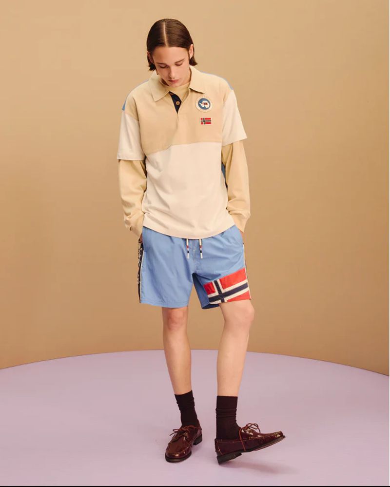Man in colour-blocked polo and blue shorts with a Norwegian flag detail, standing in a studio with a curved background.