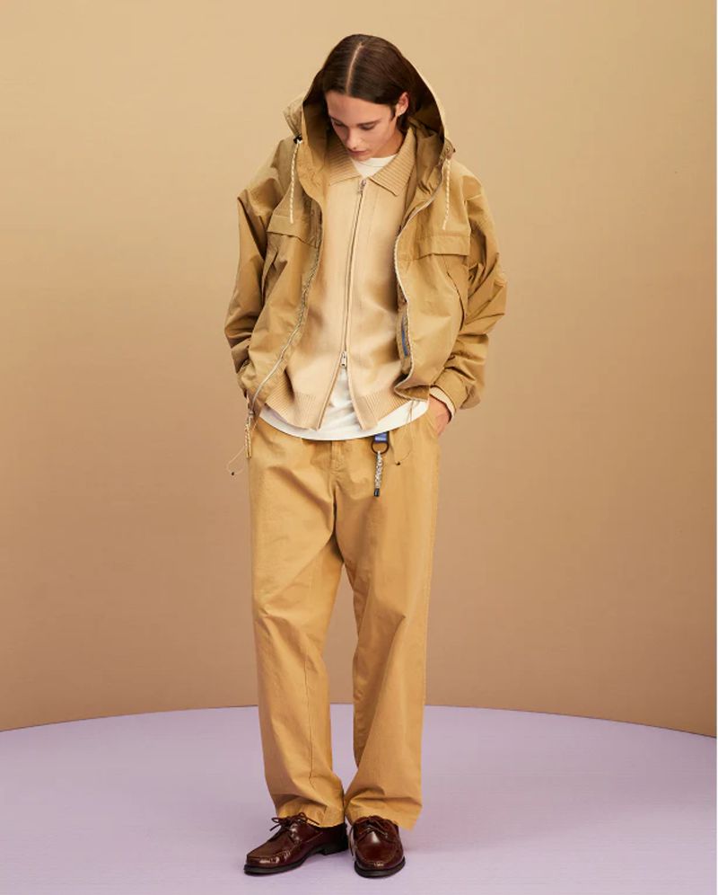Studio shot of a long-haired young man in a beige hooded jacket, zip shirt, and khaki pants with leather shoes against a neutral backdrop.