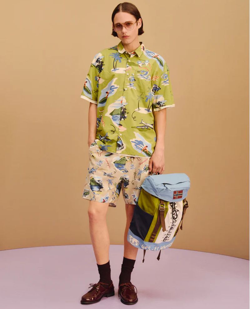 Man in tropical print shirt and shorts holding a backpack, posing in a studio with a neutral backdrop and studio lighting.