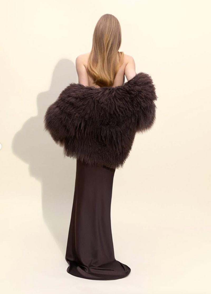 Back view of a fashion model posing in a long brown dress and draped in a textured, dark brown fur coat against a light backdrop.