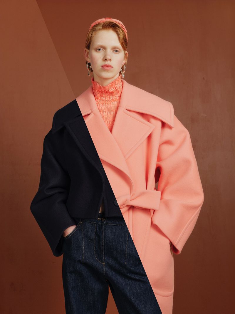 Fashionable woman posing in a black and pink color-blocked coat with jeans, hoop earrings, and a matching headband against a brown backdrop.