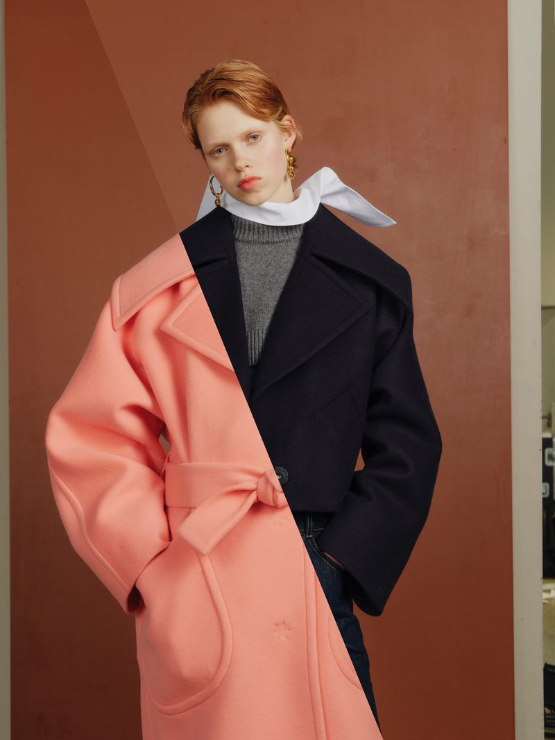 Fashionable woman in studio with split-design coat—peach and navy, gray sweater, scarf, gold earrings; modern and chic.