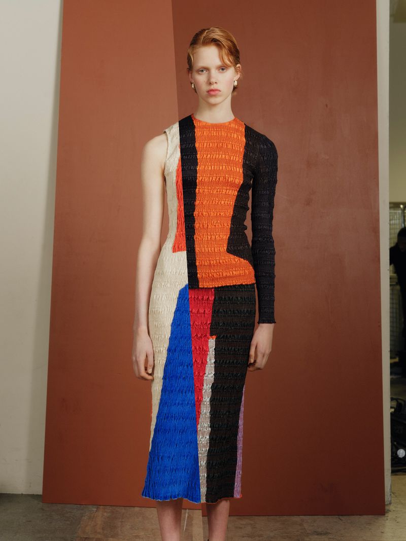 A fair-skinned fashion model stands in a studio wearing a geometrically patterned, multicolored dress, posing against a brown backdrop.