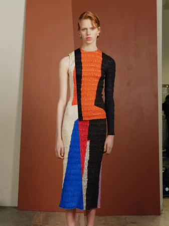A fair-skinned fashion model stands in a studio wearing a geometrically patterned, multicolored dress, posing against a brown backdrop.