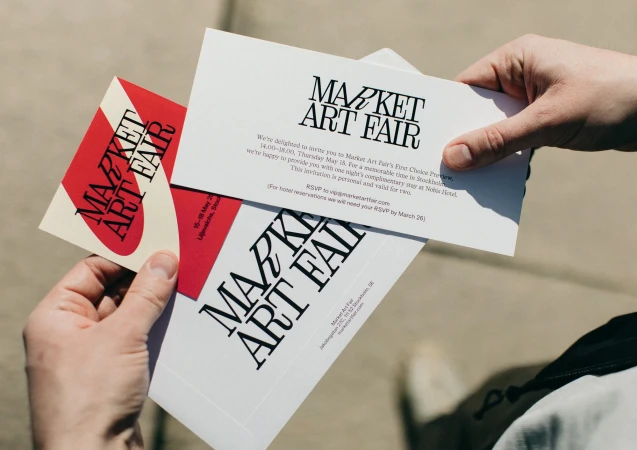 A person holds three 'Market Art Fair' invitation cards. The cards feature bold typography, and the hand is visible against a neutral background.